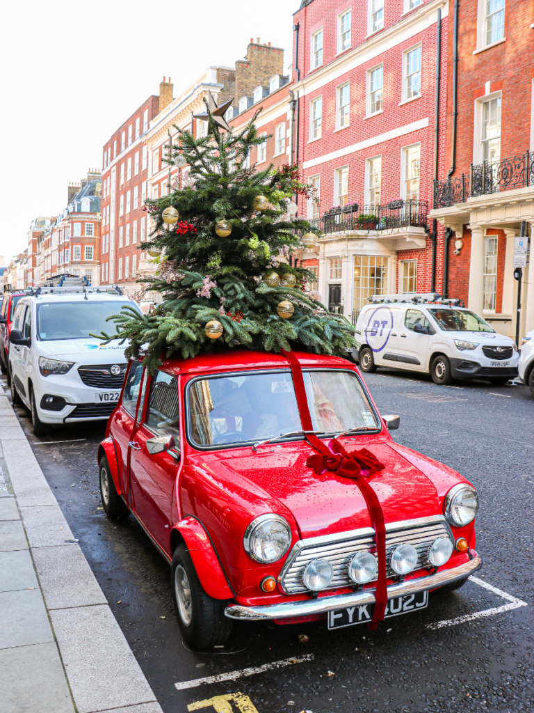 an old red mini with a large red bow on the bonnet. A small decorated Christmas tree sits on the roof, and the inside is stuffed with presents