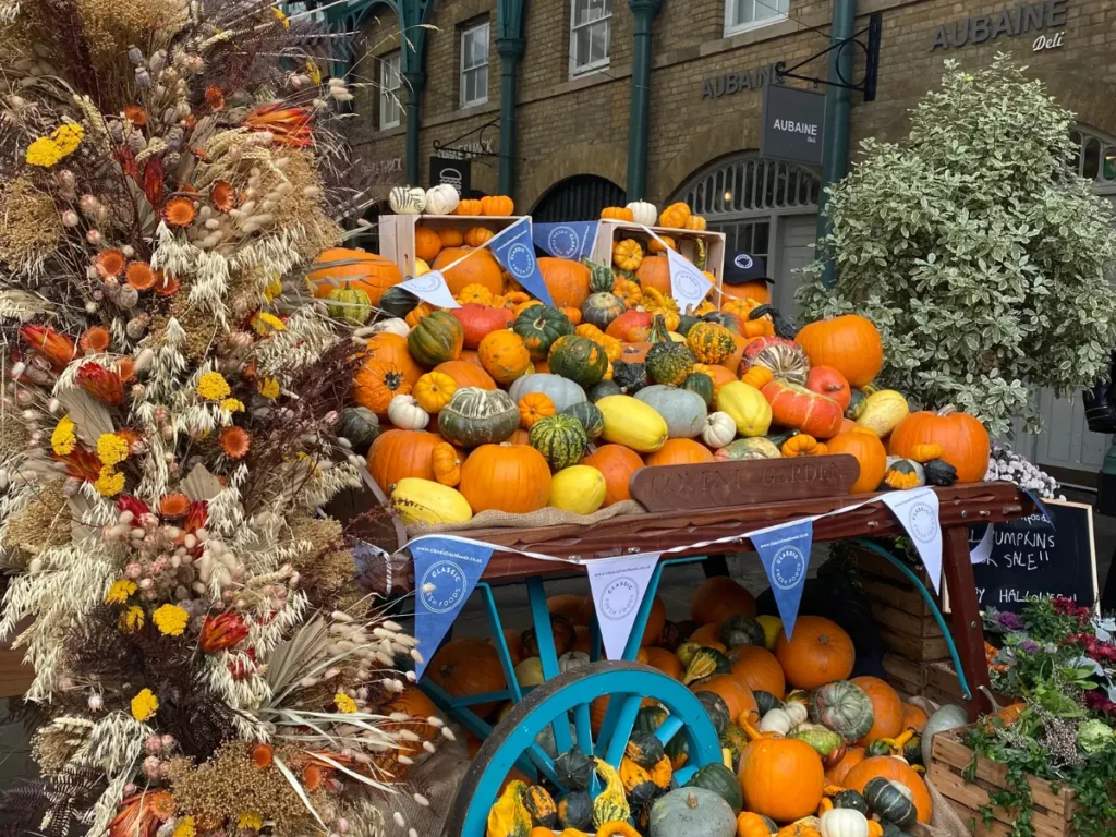 A large pile of pumpkins and ghourds in orange, yellow, white and green on a wooden trolley on blue metal wheels for Halloween