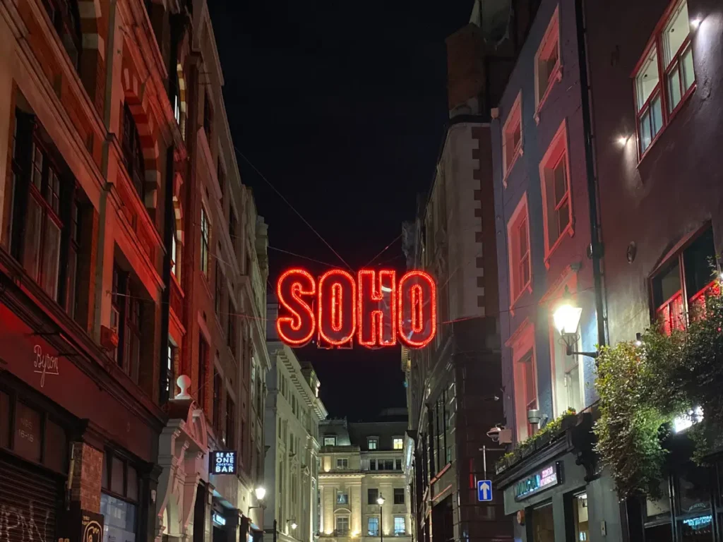 A large neon red sign that says 'SOHO' strung between two buildings at night