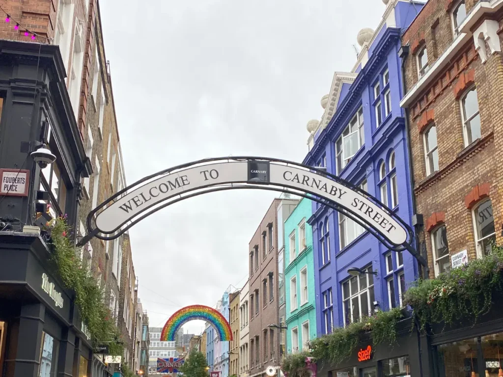 A white arched sign that says 'Welcome to Carnaby Street' in black writing. Hung between two buildings with a shopping street behind